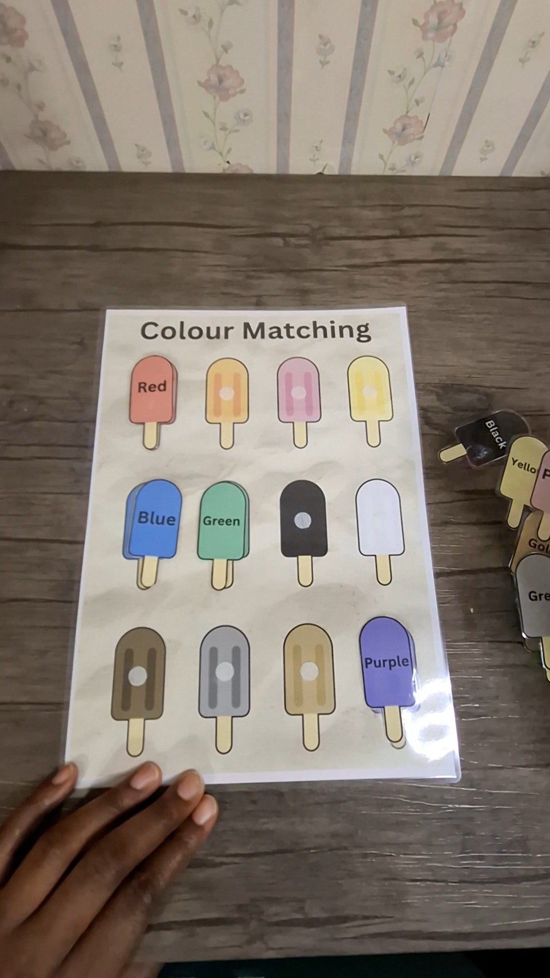 A "Colour Matching" activity featuring twelve illustrated ice lollies in various colours, including red, orange, pink, yellow, blue, green, black, white, brown, grey, gold, and purple. The ice lollies are arranged in a grid against a sandy beach background, with the title "Colour Matching" displayed in bold black text at the top.